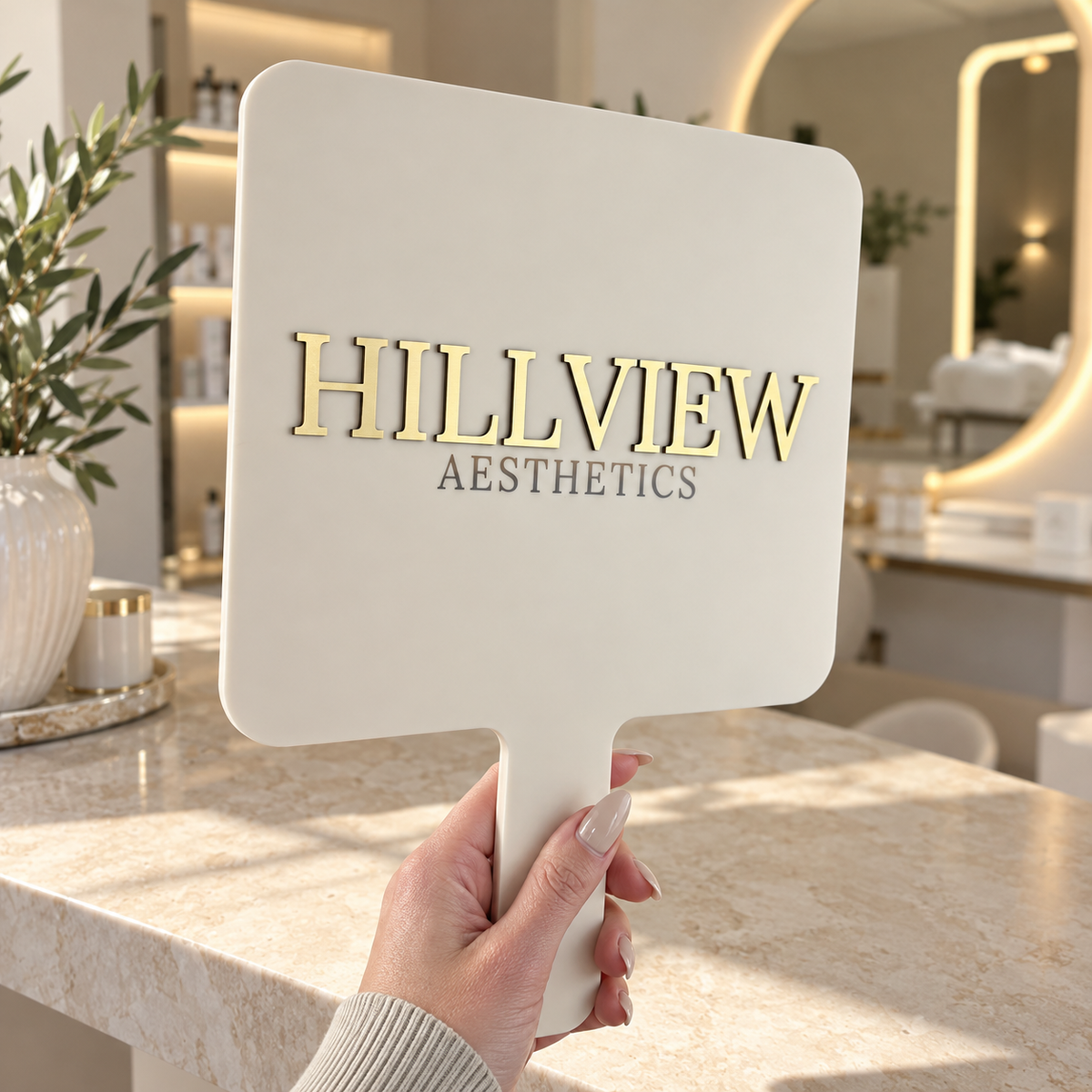 Custom cream handheld mirror with raised gold Hillview Aesthetics logo, photographed in a modern luxury beauty clinic.