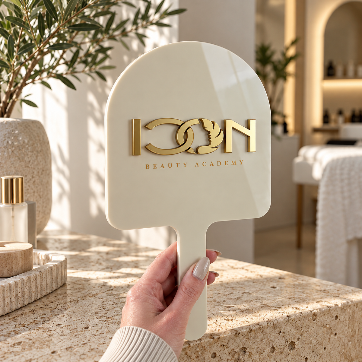 Custom cream handheld mirror with raised gold Icon Beauty Academy logo, styled in a modern beauty studio setting.