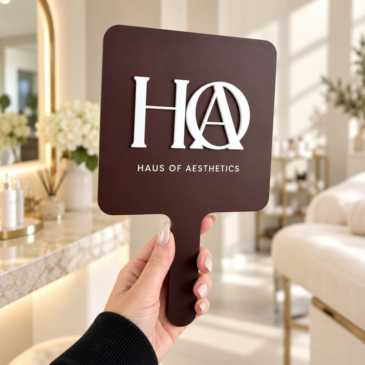 Custom branded handheld mirror with raised 3D acrylic logo for Haus of Aesthetics, photographed inside a modern beauty clinic.