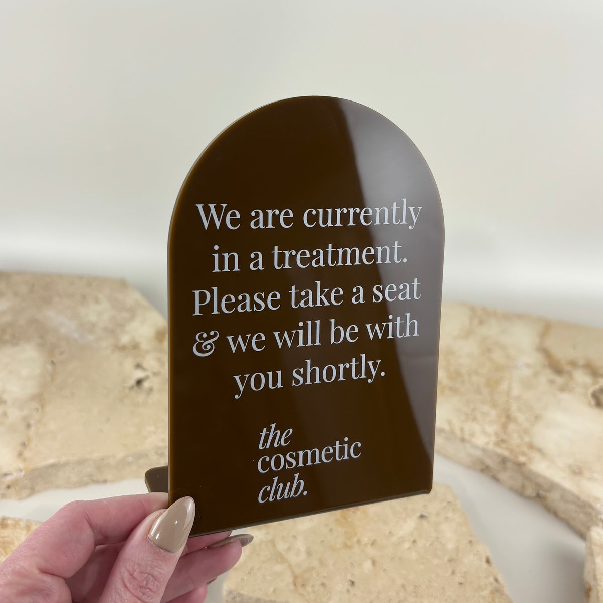 Custom glossy brown acrylic arch countertop sign with white printed treatment in progress message and The Cosmetic Club branding for a beauty clinic.