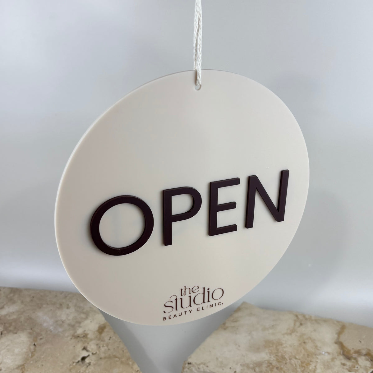 Custom round cream acrylic OPEN sign for The Studio Beauty Clinic, featuring raised dark brown 3D lettering, printed business logo and white hanging rope.