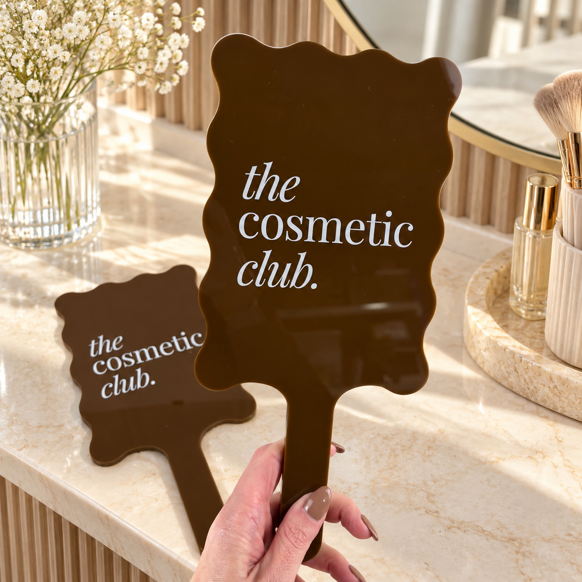 Custom brown wavy handheld mirror with white Cosmetic Club branding, styled on a modern beauty vanity with warm natural lighting.