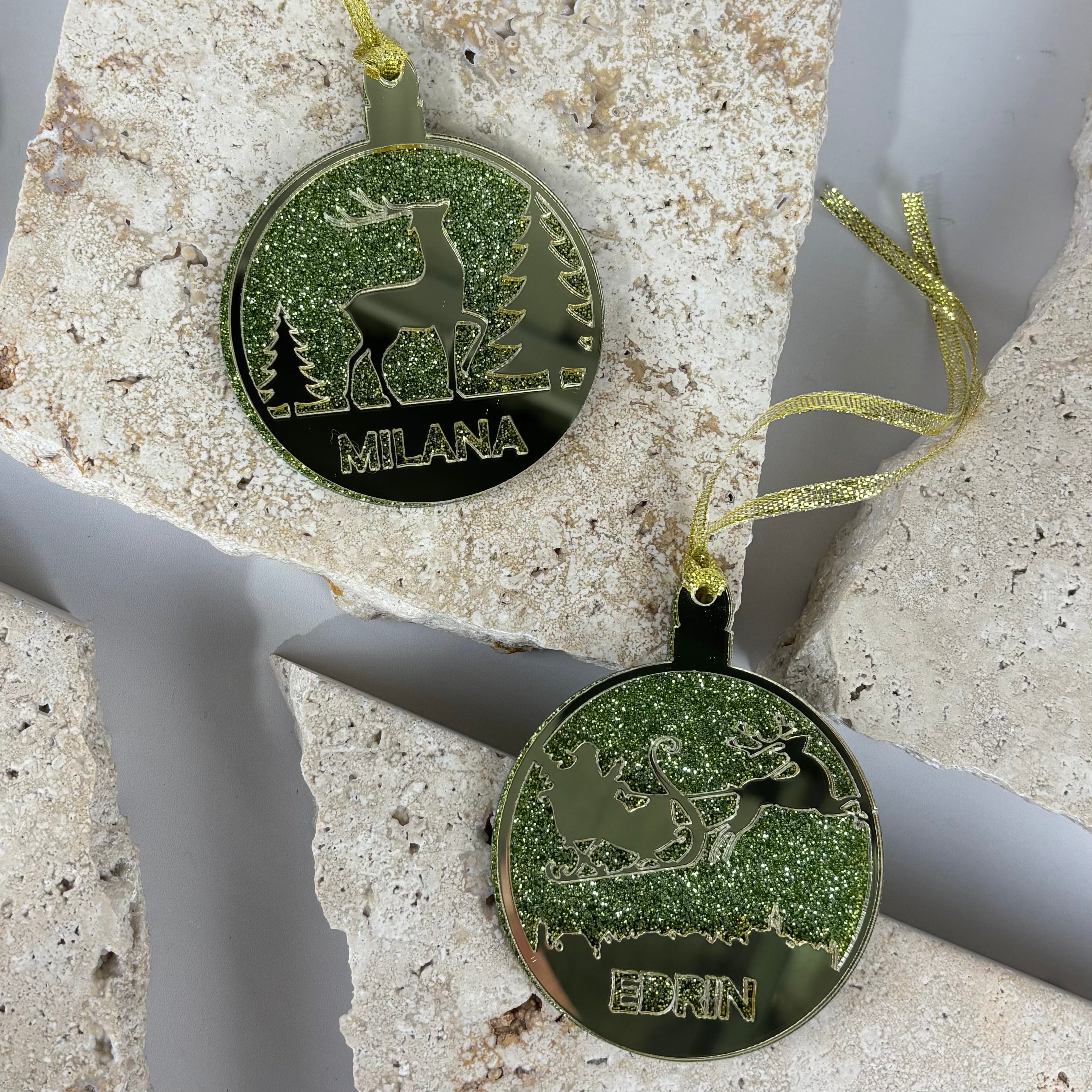 Personalised green glitter acrylic Christmas baubles with engraved reindeer and sleigh design, custom name ornaments “Milana” and “Edin”, mirrored acrylic festive tree decorations.