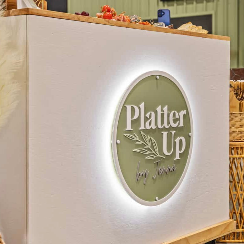 Custom round backlit business sign installed on a white catering counter for Platter Up by Jenna, featuring 3D acrylic lettering on a sage green backing, laser cut and illuminated for event display.