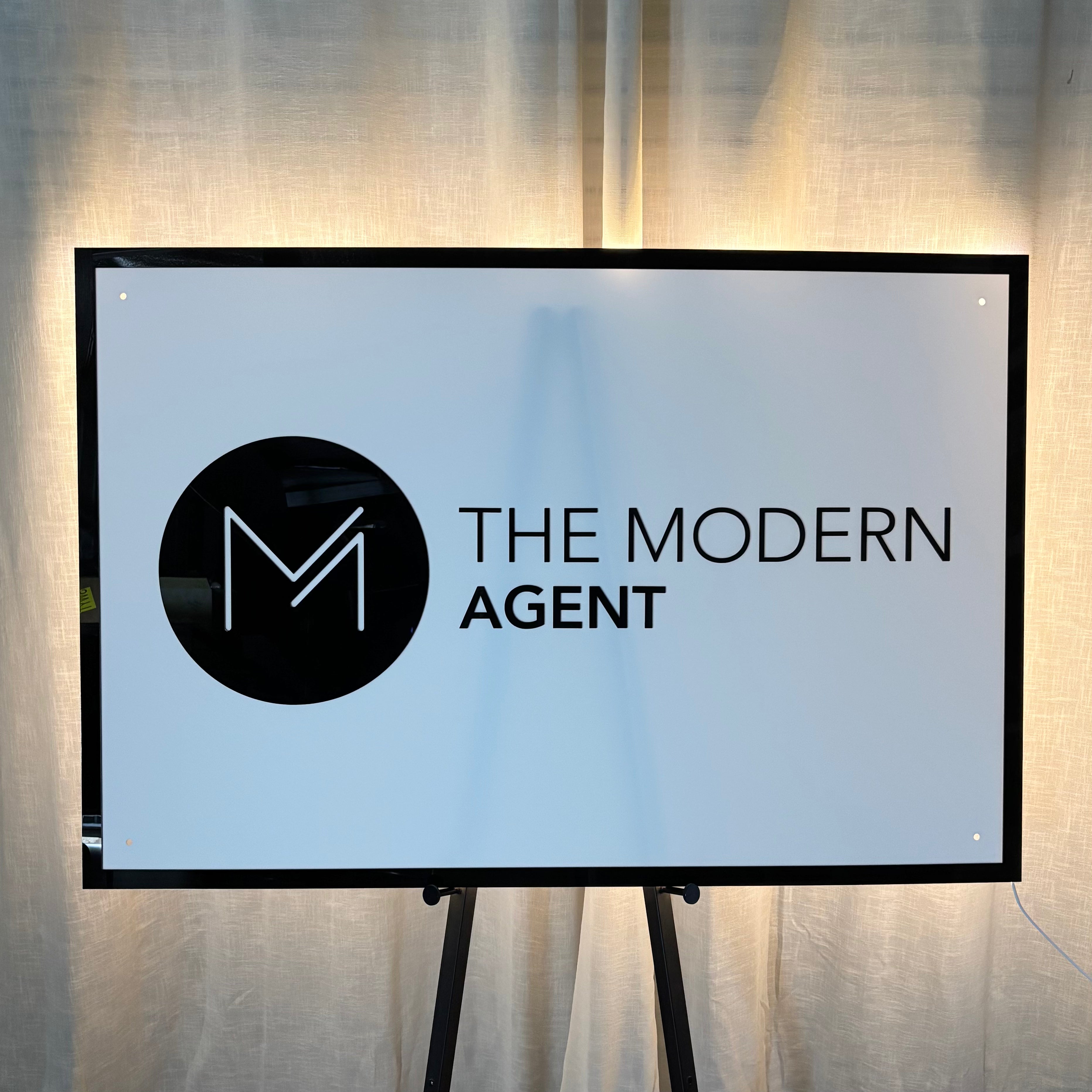 Backlit acrylic business sign for The Modern Agent, featuring a white illuminated panel with black circular logo and text, mounted on a display stand as a premium LED signage solution for real estate offices and reception areas.