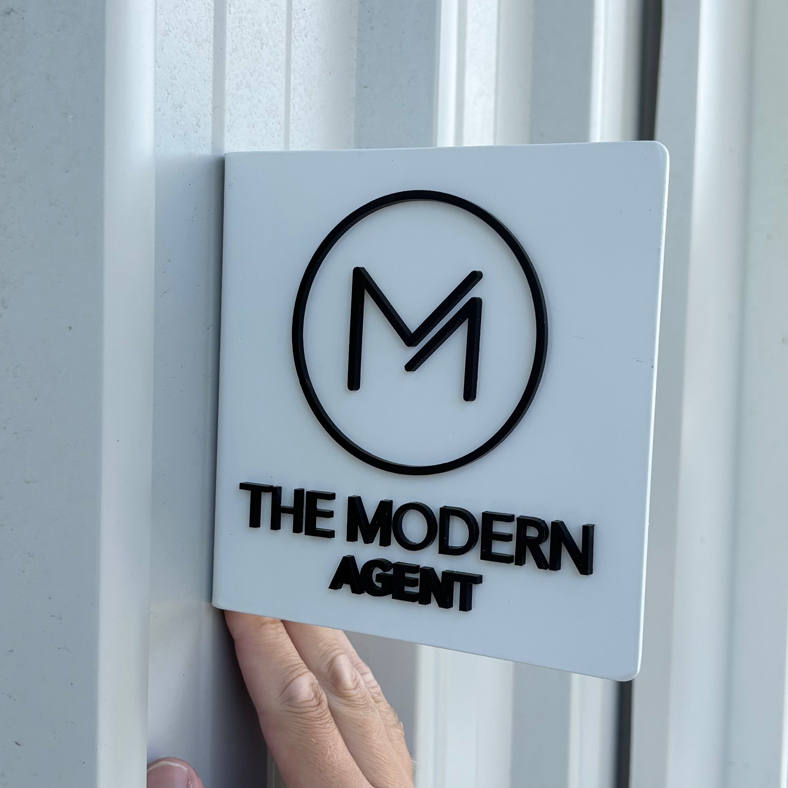 Double-sided acrylic blade sign for The Modern Agent, featuring raised black logo and lettering on a matte white square panel, mounted perpendicular to an exterior wall as a modern real estate office blade sign.
