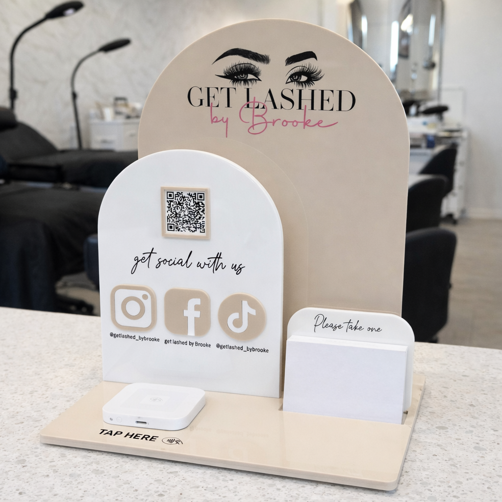 Custom acrylic NFC business stand for lash extensions salon displayed on a reception counter, featuring Get Lashed by Brooke branding, social media icons, QR code, business card holder and phone charging base