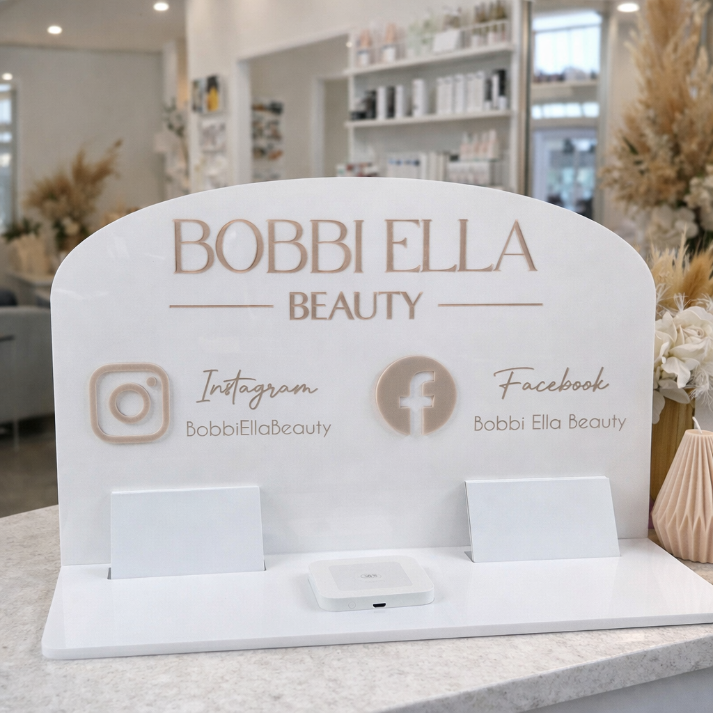 Custom white acrylic NFC business stand for beauty salon displayed on a reception desk, featuring Bobbi Ella Beauty branding, Instagram and Facebook details, business card holders and square reader device for tap payments