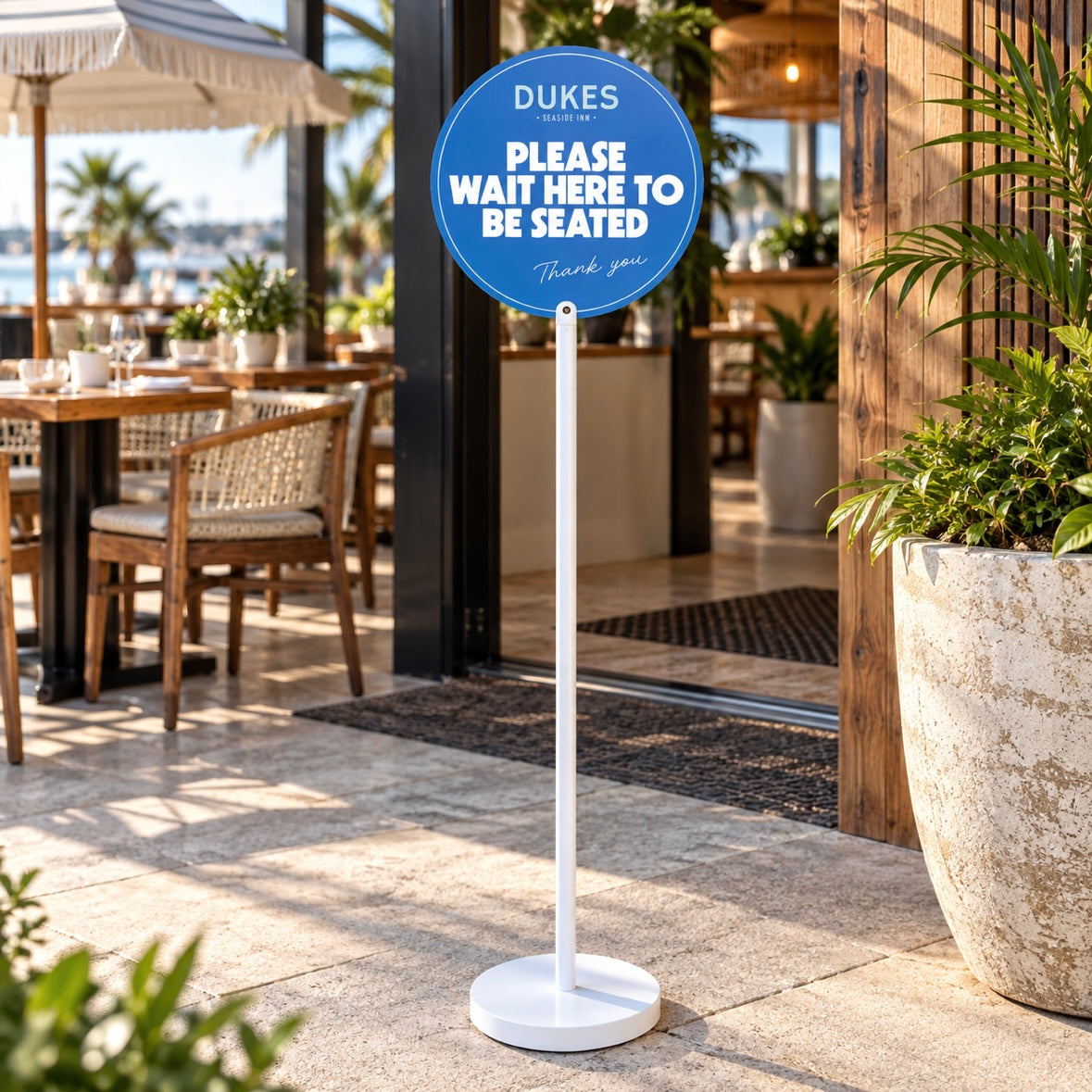 Freestanding restaurant wait sign with round blue acrylic face and white stand at a modern seaside restaurant entrance.