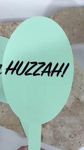 Close up of green acrylic prop paddle with bold Huzzah text. Video 